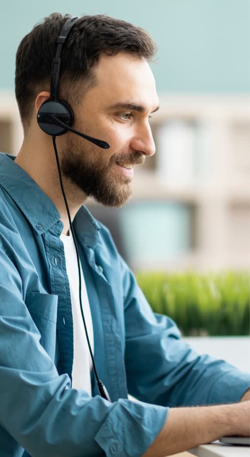 Der Mann trägt ein Headset und arbeitet an einem Laptop in einem modernen Büro. Mit einem konzentrierten Blick scheint er eine Aufgabe zu bearbeiten, möglicherweise im Kundenservice oder in der technischen Unterstützung. Der Raum ist gut beleuchtet, und im Hintergrund sind Pflanzen zu sehen, die eine angenehme und produktive Arbeitsatmosphäre schaffen.