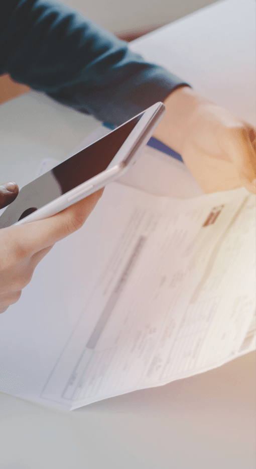 Eine Person sitzt an einem Tisch, hält ein Smartphone in der einen Hand, während sie mit der anderen Hand ein Dokument mit gedruckten Details hält. Die Szene deutet auf eine geschäftliche oder finanzielle Aufgabe hin, wobei ein Taschenrechner und einige Unterlagen auf dem Tisch sichtbar sind. Das Bild hebt die Nutzung mobiler Technologie zur Verwaltung oder Bearbeitung von Dokumenten hervor.