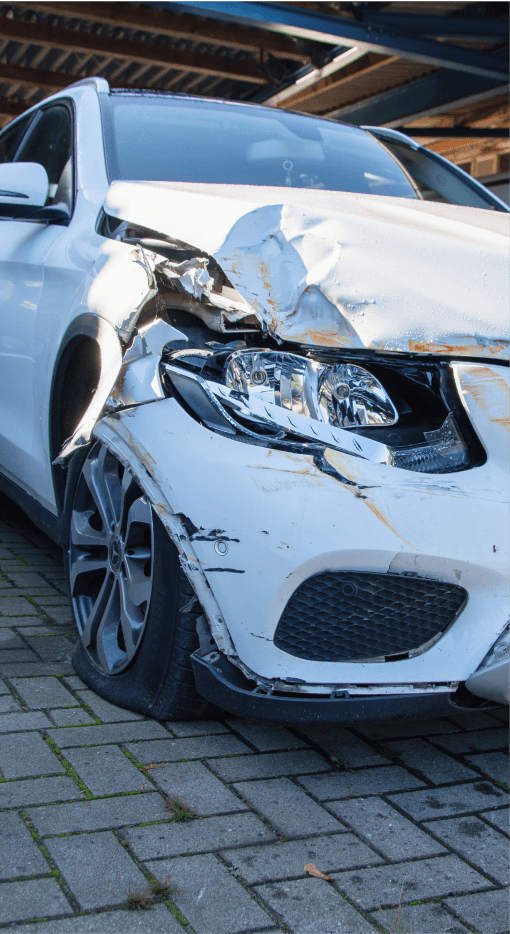 Zu sehen ist ein beschädigtes Fahrzeug, das einen erheblichen Schaden an der vorderen linken Seite aufweist. Der Stoßfänger und die Karosserie sind stark verbeult, und der Reifen ist platt. Es scheint sich um einen Unfall mit frontalen Schäden zu handeln, wobei das Fahrzeug auf einem gepflasterten Untergrund geparkt ist. Die Beleuchtung und der Kühlerbereich des Fahrzeugs sind ebenfalls betroffen.