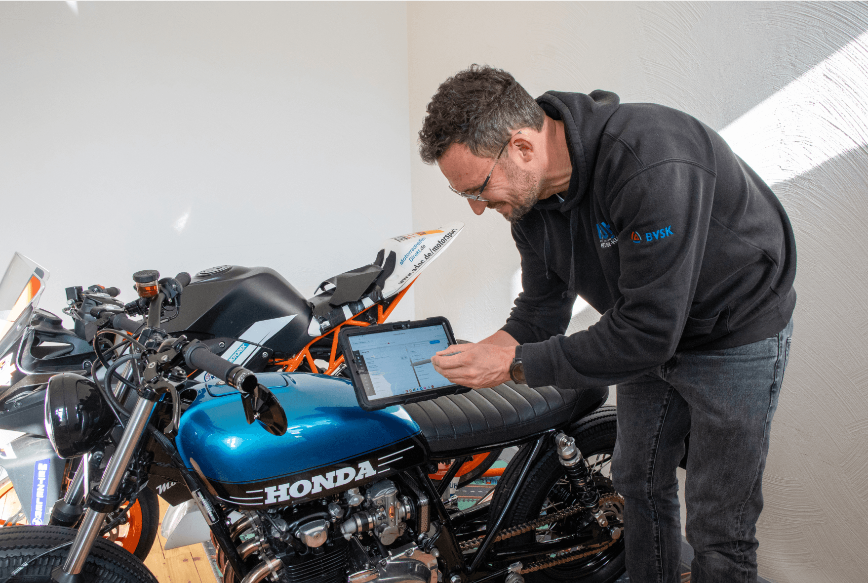 Das Bild zeigt einen Kfz-Sachverständigen, der ein Tablet in der Hand hält und auf die CombiPlus Online Software zugreift, um einen Schadenfall zu bearbeiten. Er steht in einer Werkstatt und ist vor einer Honda-Motorrad mit einer auffälligen blauen Tankfarbe. Der Sachverständige trägt ein schwarzes Hoodie mit dem BVSK-Logo und hat eine konzentrierte Haltung, während er das Tablet bedient. Das Bild vermittelt den Einsatz der modernen Software für die mobile Schadenaufnahme und -bearbeitung, um die Prozesse effizient und schnell abzuwickeln.