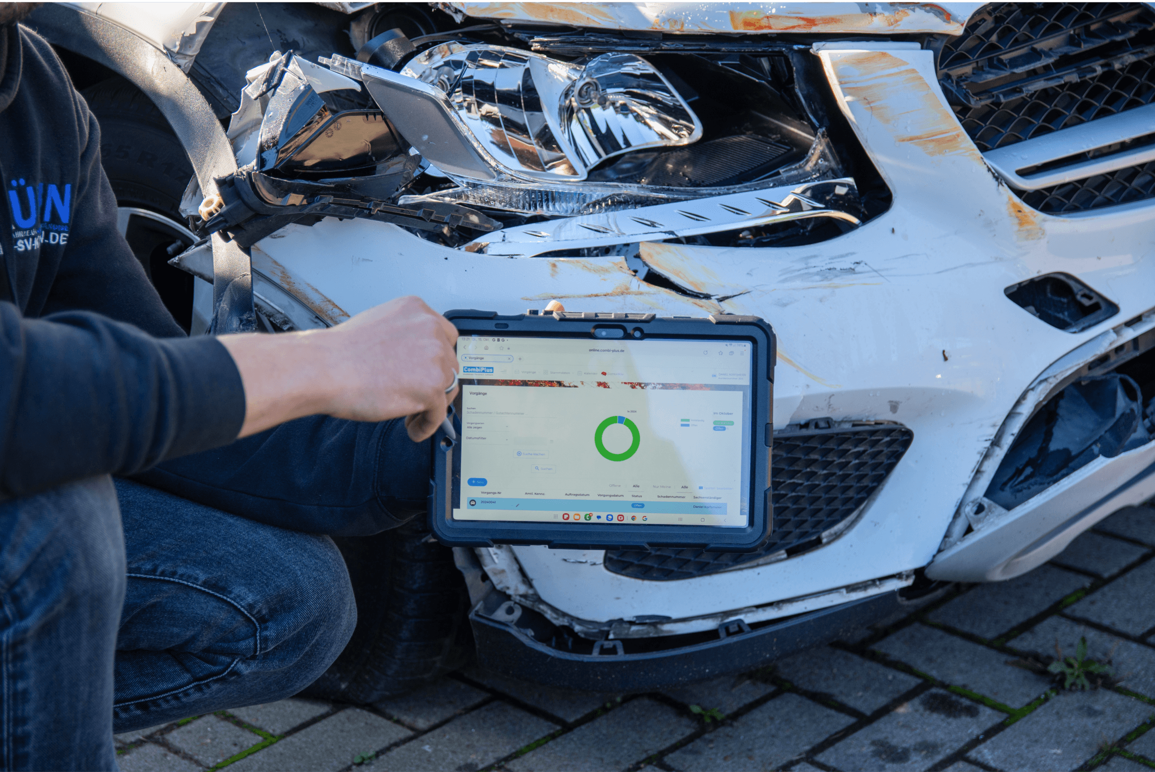 Das Bild zeigt eine Person, die ein Tablet mit der myConnect-Software vor einem beschädigten Fahrzeug hält. Das Fahrzeug hat einen erheblichen Frontschaden. Auf dem Tablet-Bildschirm ist eine grüne Anzeige sichtbar, die möglicherweise den Status eines Schadenfalls darstellt. Die Person, die das Tablet hält, ist in einem schwarzen Sweatshirt gekleidet und kniet neben dem Fahrzeug. Der Hintergrund zeigt Asphalt und einen Teil des beschädigten Autos.