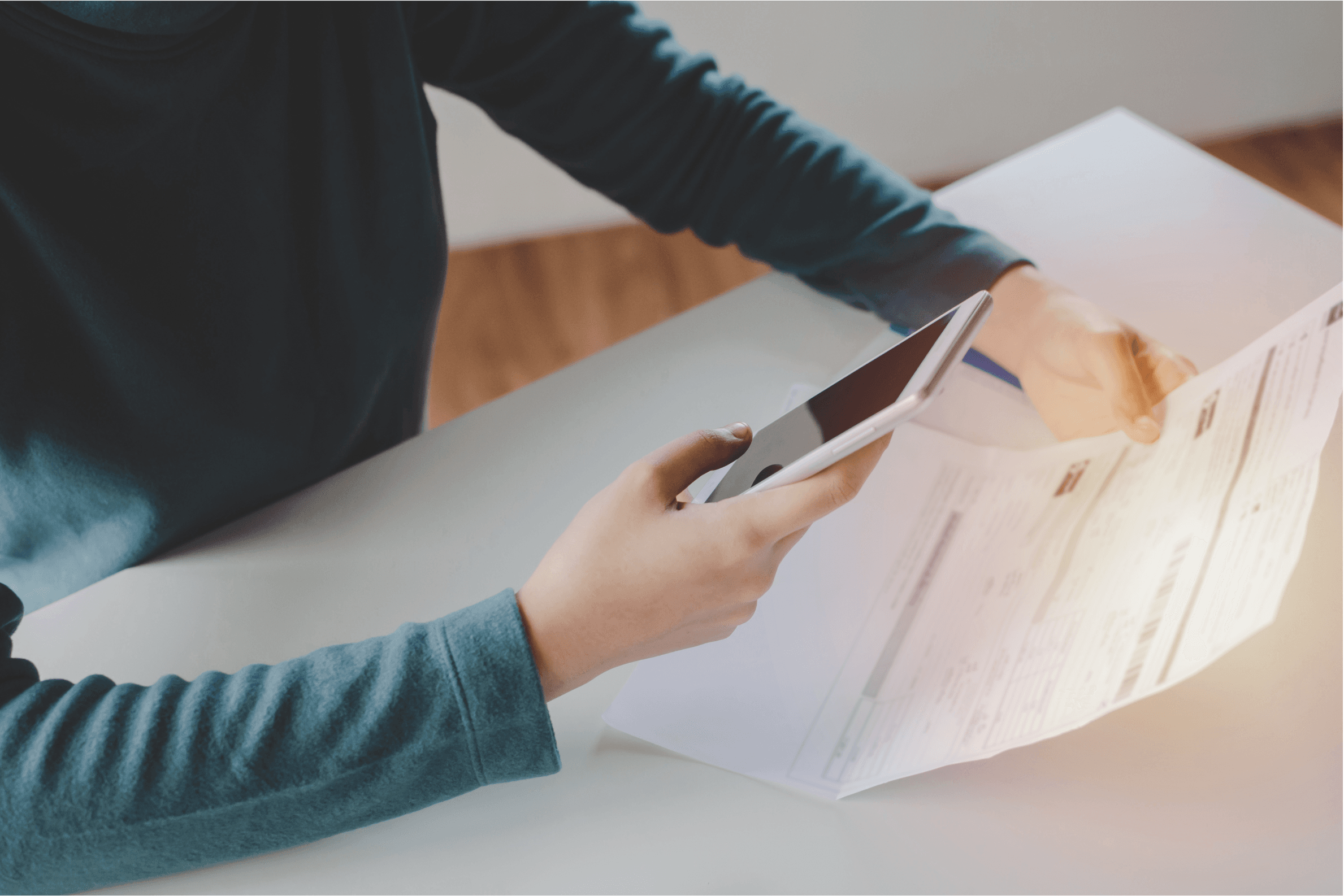 Das Bild zeigt eine Person, die mit einer Hand ein Smartphone hält und mit der anderen Hand ein Dokument auf einem Tisch betrachtet. Der Hintergrund ist unscharf und zeigt einen minimalistischen Raum. Das Dokument, das in der Hand gehalten wird, scheint eine Rechnung zu sein, aber der Text darauf ist unscharf. Die Person ist in einem blauen Oberteil gekleidet und sitzt an einem Tisch, auf dem auch weiteres Papier zu sehen ist.