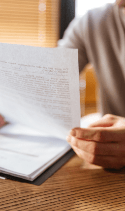 Das Bild zeigt eine Person, die in einem Büro an einem Tisch sitzt und ein Dokument in den Händen hält. Der Fokus liegt auf der Person und den Dokumenten, während der Hintergrund unscharf bleibt. Die Person scheint das Dokument zu lesen oder durchzublättern, wobei die Beleuchtung und der Holztisch eine angenehme Atmosphäre schaffen.