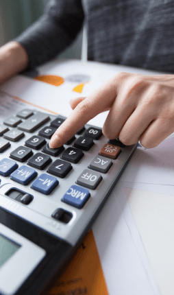 Das Bild zeigt eine nahaufgenommene Hand einer Person, die mit einem Taschenrechner arbeitet. Die Person tippt auf die Tasten des Rechners, während der Hintergrund unscharf ist und einige Dokumente zu sehen sind. Es scheint, dass die Person gerade eine Berechnung durchführt.