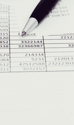 Das Bild zeigt einen Schreibtisch mit einer Tabelle und einem Kugelschreiber. Die Tabelle enthält Zahlen, die möglicherweise finanzielle oder buchhalterische Daten darstellen. Der Kugelschreiber liegt auf der Tabelle, was darauf hinweist, dass jemand gerade dabei ist, die Daten zu überprüfen oder zu notieren. Der Fokus des Bildes liegt auf einem Abschnitt der Tabelle und dem Stift.