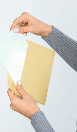 Das Bild zeigt eine Person, die ein Umschlag mit einem Blatt Papier in den Händen hält. Die Person scheint den Umschlag zu öffnen, um den Inhalt herauszunehmen. Das Papier im Umschlag ist unbeschriftet, was darauf hindeutet, dass es möglicherweise Platz für Notizen oder Dokumente bietet. Die Person trägt einen grauen Pullover, und die Handlung des Öffnens des Umschlags wirkt professionell und ordentlich.