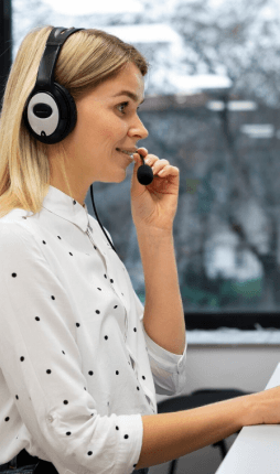 Das Bild zeigt eine Frau, die in einem Callcenter oder Büro arbeitet. Sie trägt Kopfhörer mit einem Mikrofon und spricht möglicherweise in ein Gespräch oder eine Besprechung. Ihre Mimik deutet auf eine konzentrierte Kommunikation hin, und der hintergrund ist leicht verschwommen, was auf eine professionelle Arbeitsumgebung hinweist. Sie trägt ein weißes Hemd mit Punkten, was einen gepflegten, professionellen Eindruck vermittelt. Sie repräsentiert unseren CombiSupport