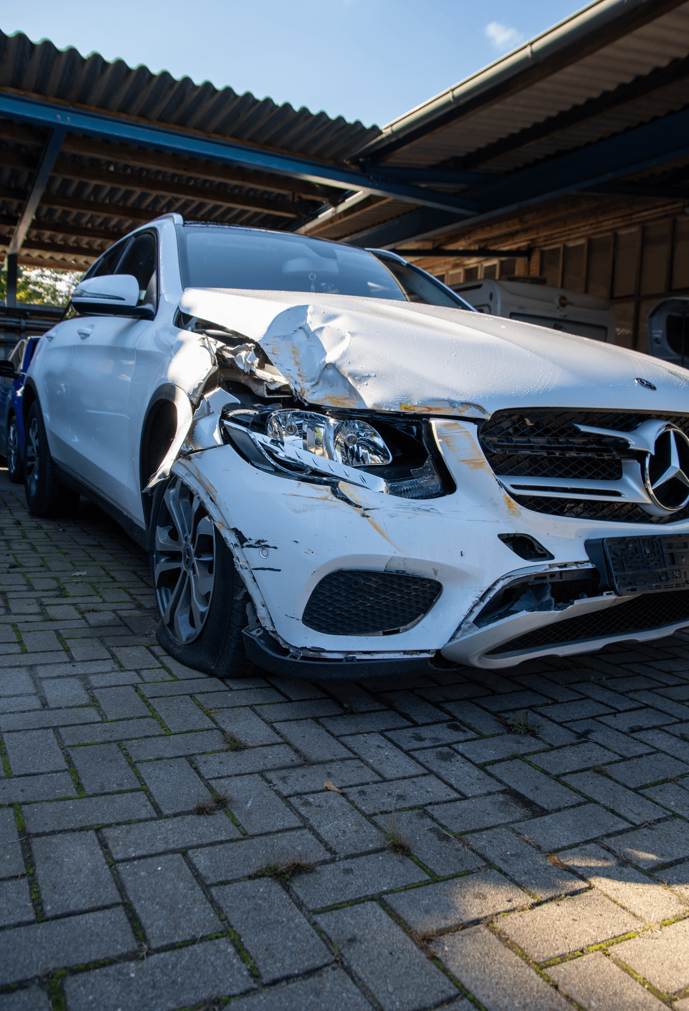 Das Bild zeigt ein stark beschädigtes weißes Mercedes-Benz-Fahrzeug mit erheblichen Schäden an der Vorderseite. Der vordere Stoßfänger und die Fahrzeugfront sind verbeult, und der Reifen auf der Fahrerseite ist abgeflacht, was auf einen möglichen Unfall oder eine Kollision hindeutet. Im Hintergrund ist eine Werkstattstruktur sichtbar, was darauf hindeutet, dass das Fahrzeug zur Schadensbewertung oder Reparatur vorbereitet wird.
