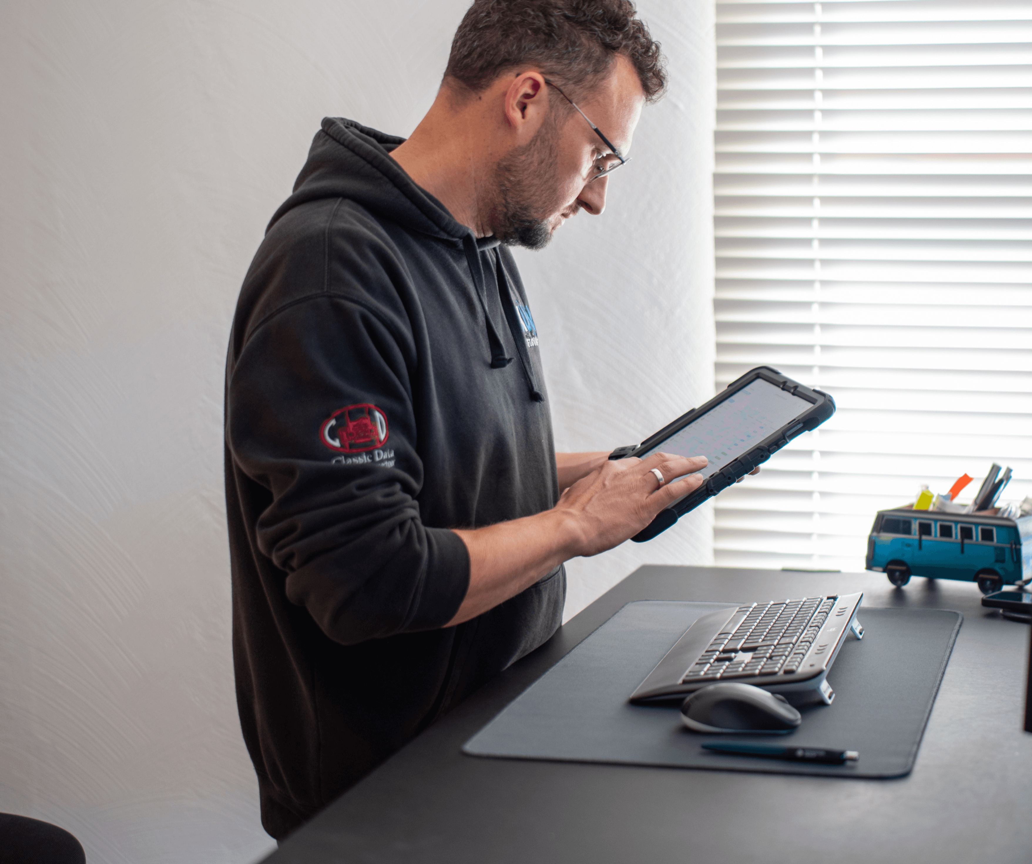 Ein Kfz-Sachverständiger hält ein Tablet in der Hand und schaut auf den Bildschirm. Auf dem Tablet ist die CombiPlus Online Software sichtbar, die für die mobile Schadenaufnahme und die Bearbeitung von Gutachten verwendet wird. Der Sachverständige befindet sich an einem Schreibtisch, der mit einer Tastatur, einer Maus und Schreibutensilien ausgestattet ist. Auf dem Tisch steht ein kleines Modell eines VW-Busses, was auf die Arbeit des Sachverständigen im Bereich Kfz-Gutachten hinweist. Er trägt einen schwarzen Pullover mit dem Logo Classic Data auf dem Ärmel. Die Umgebung ist modern und gut beleuchtet, mit einem Fenster im Hintergrund, das Tageslicht hereinlässt. Das Bild vermittelt eine produktive und technologieorientierte Arbeitsumgebung, in der CombiPlus Online als Werkzeug zur effizienten Schadenbearbeitung genutzt wird.