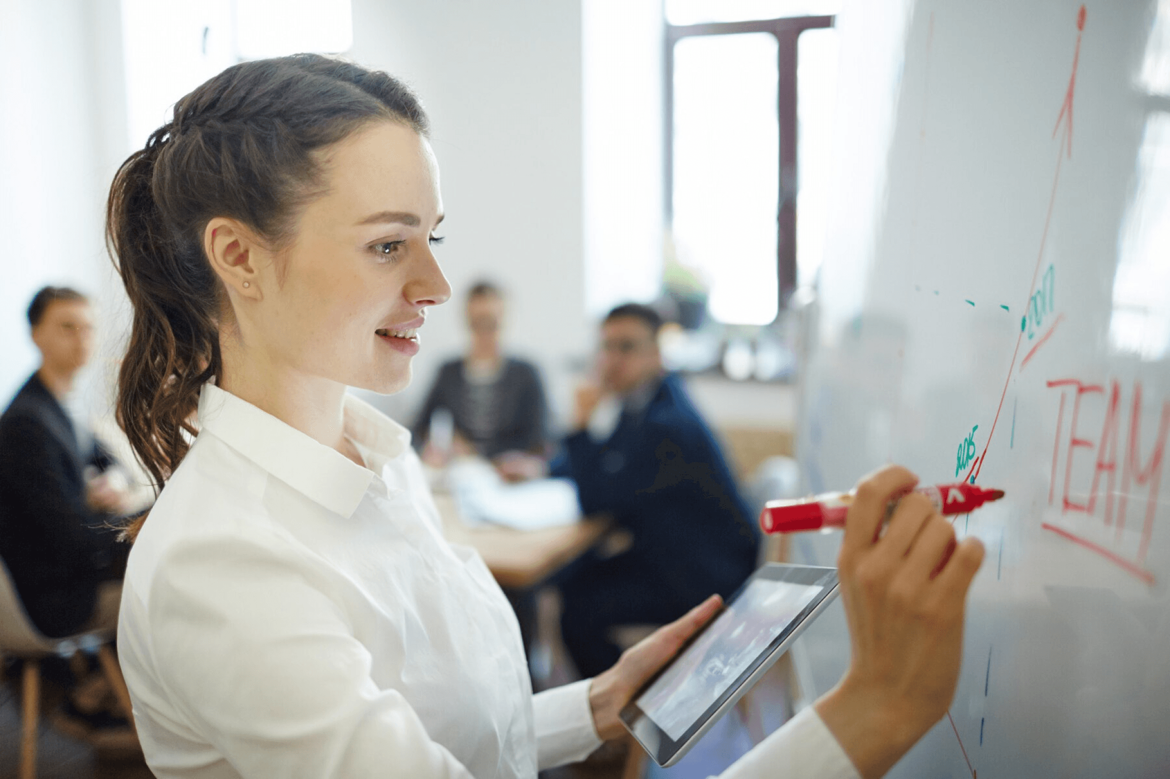 Das Bild zeigt eine junge Frau, die in einem Konferenzraum vor einem Whiteboard steht und mit einem roten Marker darauf schreibt. In ihrer linken Hand hält sie ein Tablet, während sie mit der rechten Hand den Marker führt. Auf dem Whiteboard sind Grafiken und Begriffe wie "TEAM" sowie ein Diagramm zu sehen, das Wachstum oder Fortschritte darstellt. Im Hintergrund sind mehrere Personen in einem Meeting zu sehen, die aufmerksam zuhören oder mitarbeiten. Das Bild vermittelt eine professionelle und kollaborative Arbeitsatmosphäre.