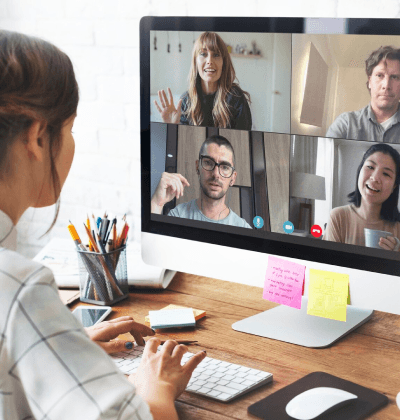 Das Bild zeigt eine Person, die an einem Schreibtisch arbeitet und an einem virtuellen Meeting teilnimmt. Auf dem Bildschirm vor ihr sind vier weitere Teilnehmer zu sehen, die in einem Video-Call miteinander sprechen. Die Teilnehmer lächeln oder winken, was eine freundliche und interaktive Atmosphäre vermittelt. Auf dem Schreibtisch sind verschiedene Büroartikel wie Stifte, Notizblätter und Post-its zu sehen, die auf eine produktive Arbeitsumgebung hinweisen. Das Bild vermittelt den Eindruck eines modernen, flexiblen Arbeitens in einem digitalen Team-Setting.