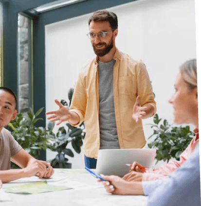 Das Bild zeigt eine Gruppe von vier Personen in einem modernen Büro, die an einem Tisch sitzen. Ein Mann steht auf und spricht, während die anderen aufmerksam zuhören. Er trägt ein lockeres Hemd und eine Brille und scheint eine Idee oder einen Vorschlag zu präsentieren. Auf dem Tisch liegen Notizen und ein Laptop. Der Raum ist gut beleuchtet, mit Pflanzen im Hintergrund, die eine angenehme Arbeitsatmosphäre schaffen. Es vermittelt einen kreativen und kollaborativen Moment in einer Besprechung oder Diskussion.