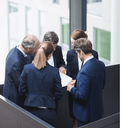 Das Bild zeigt eine Gruppe von fünf Personen in formeller Kleidung, die in einem modernen Bürogebäude zusammenstehen. Sie tragen alle Anzüge und diskutieren intensiv über ein Dokument, das eine Person in der Gruppe in den Händen hält. Die anderen scheinen die Informationen auf dem Blatt zu überprüfen oder darüber zu sprechen. Die Szene vermittelt eine professionelle Arbeitsatmosphäre, in der Teamarbeit und Zusammenarbeit im Vordergrund stehen. Das Büro im Hintergrund ist hell und modern gestaltet, mit großen Fenstern, die Tageslicht hereinlassen.