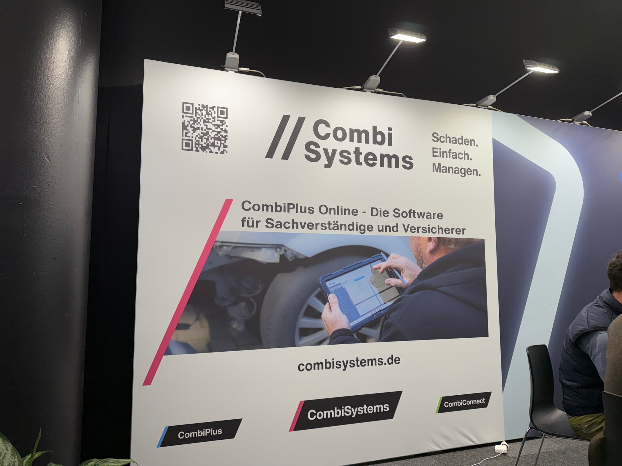 Messestand von CombiSystems auf der AutoZum in Salzburg. Der Stand zeigt ein großes Banner mit dem Logo von CombiSystems und dem Slogan 'Schaden. Einfach. Managen.' sowie die Beschreibung 'CombiPlus Online - Die Software für Sachverständige und Versicherer'. Auf dem Banner ist ein Bild eines Tablets mit der Software abgebildet. Oben links befindet sich ein QR-Code, und unten sind die Produkte 'CombiPlus' und 'CombiConnect' dargestellt. Der Stand ist modern beleuchtet.