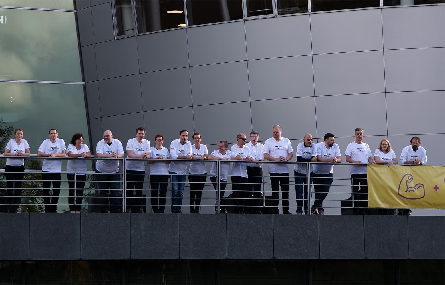 Das CombiTeam von CombiSystems steht auf einem Balkon der Gläsernen Manufaktur in Dresden. Alle tragen weiße T-Shirts mit dem Logo von CombiSystems. Rechts im Bild ist ein gelbes Banner mit einem Symbol eines starken Arms und einem Pluszeichen zu sehen, das Stärke und Teamarbeit symbolisiert.