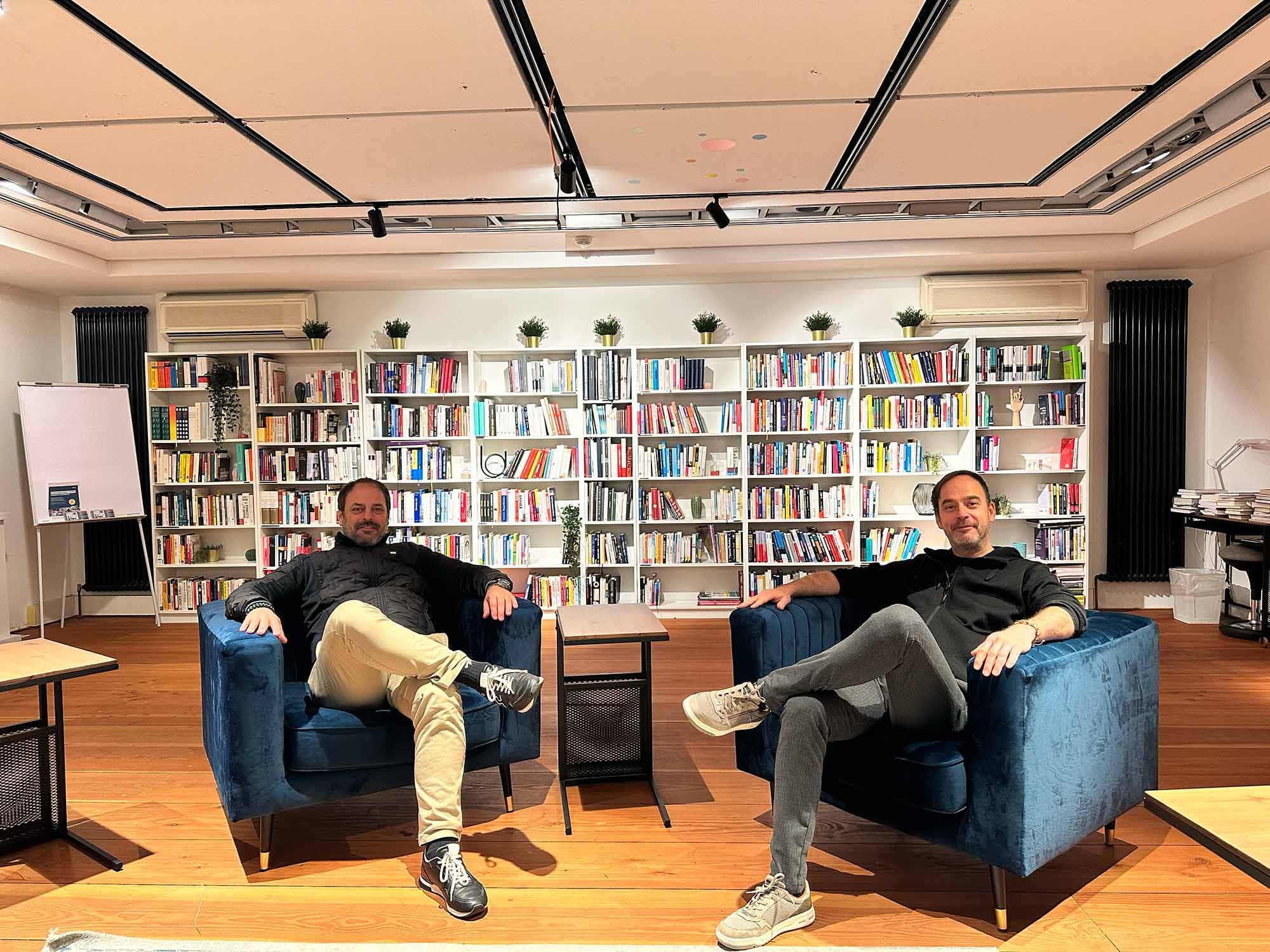 Stefan Grimm und Sebastian Grimm sitzen entspannt in blauen Sesseln in einer modernen Bibliothek mit deckenhohen weißen Bücherregalen. Die Atmosphäre im MachWerk ist gemütlich, mit Pflanzen auf den Regalen, einem Holzboden und warmem Licht.